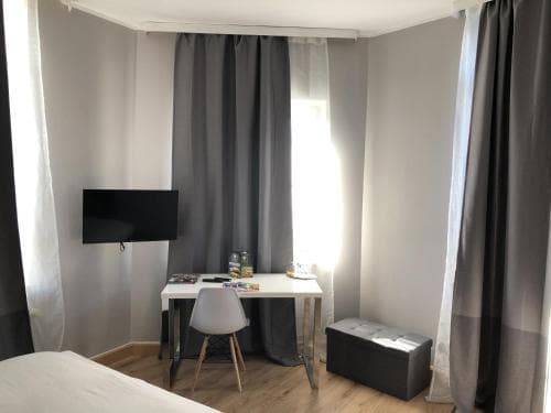 A cozy hotel room features a neatly arranged desk with a chair, a mounted TV, soft gray curtains framing a window, and a storage ottoman, ideal for travelers seeking comfort and functionality.