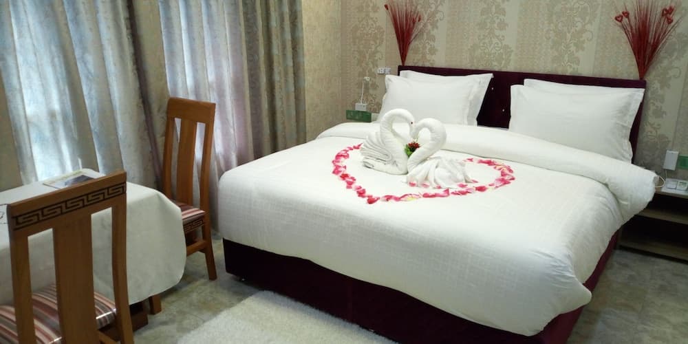 A cozy hotel room features a neatly made bed adorned with swan-shaped towels and a heart of rose petals, creating a romantic ambiance ideal for honeymooners or couples seeking a special getaway.