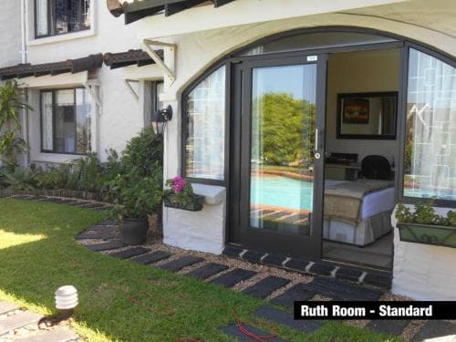 A serene Ruth Room Standard features a lush garden view through glass doors, inviting travelers to relax in a cozy bedroom with a poolside ambiance, perfect for leisure stays.