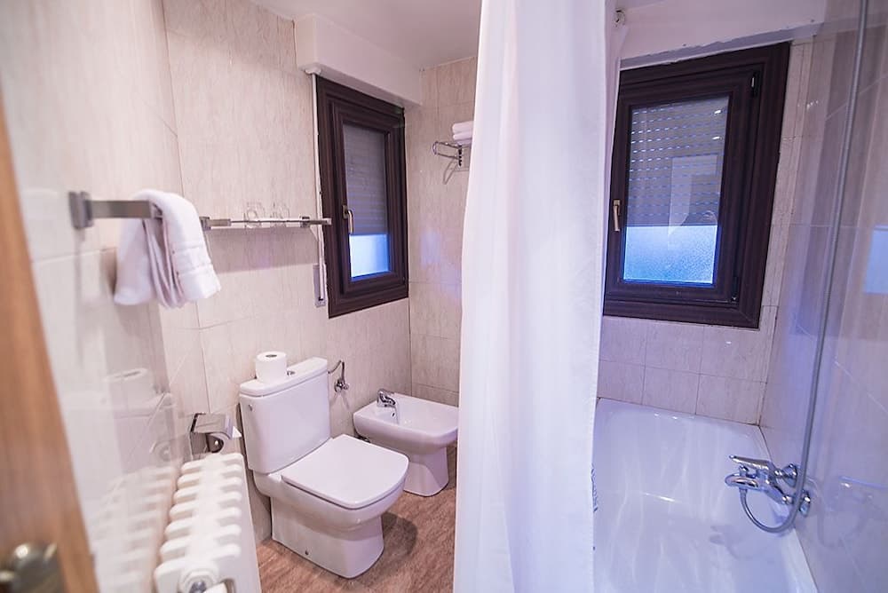A cozy hotel bathroom features tiled walls, a toilet, bidet, shower with glass doors, and windows with blinds, ideal for travelers seeking comfort and cleanliness.