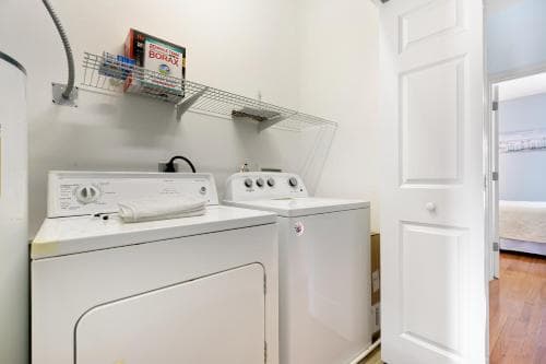 A cozy laundry room with white appliances, a metal shelving unit holding cleaning supplies, and an open door leading to a hallway, ideal for travelers seeking self-service laundry facilities in their accommodations.