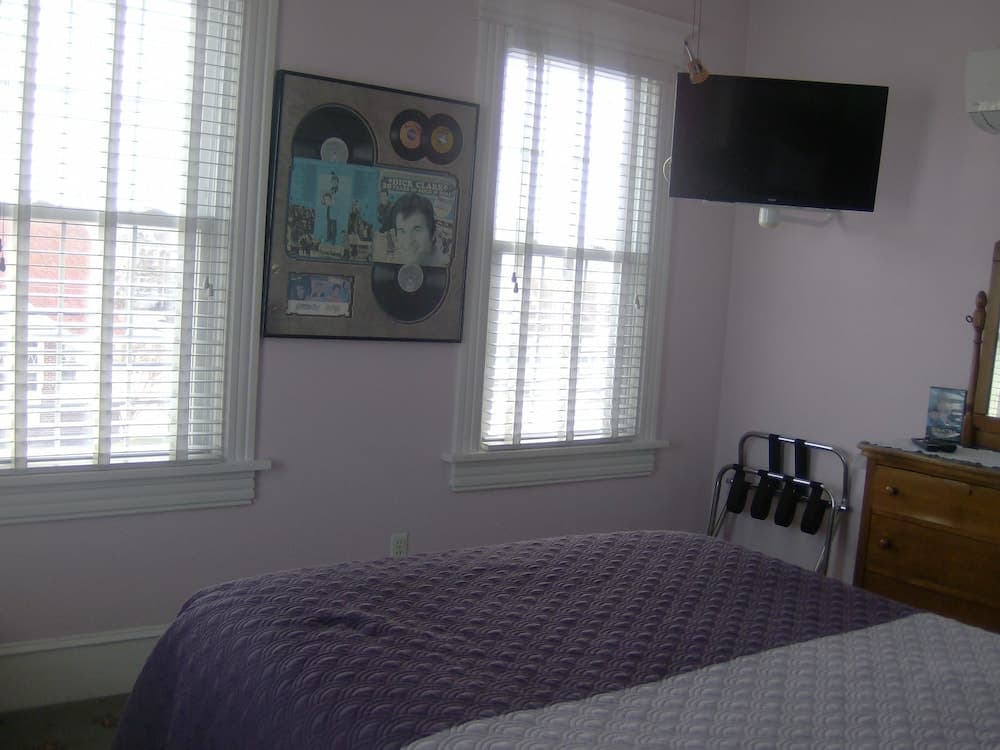 A cozy bedroom with soft purple bedding, framed vinyl records on the wall, a mounted TV, and natural light filtering through blinds, ideal for travelers seeking a comfortable and nostalgic retreat.