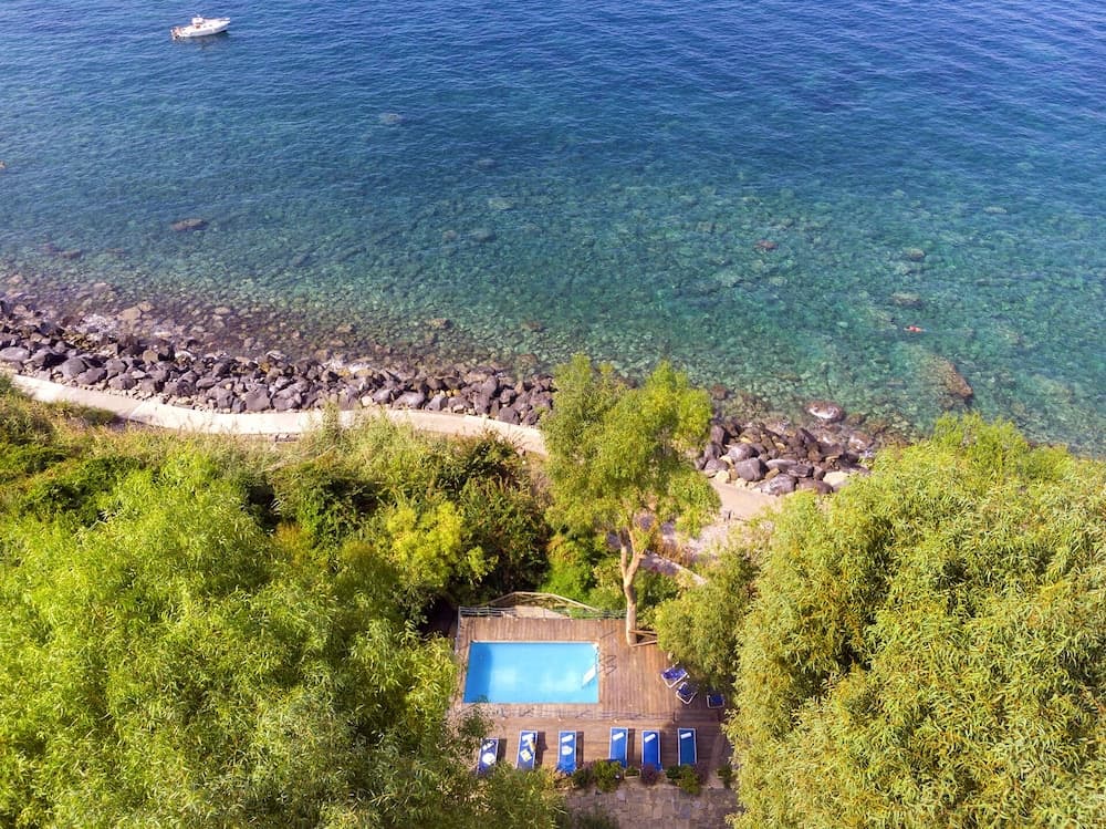 An aerial view captures a serene coastal retreat with a private pool surrounded by lush greenery, overlooking crystal-clear turquoise waters ideal for luxury beachside vacations.