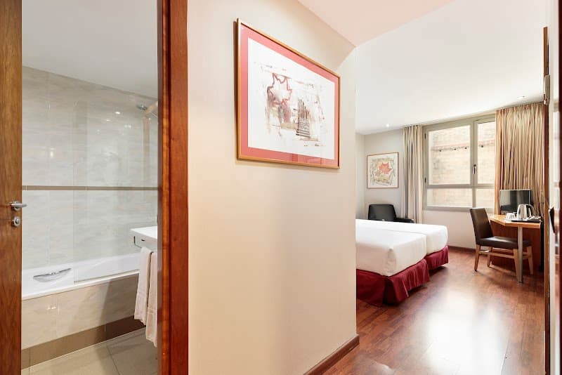 A cozy hotel room in Barcelona's Eixample district features a neatly made bed, wooden floors, modern artwork, and an adjacent bathroom, ideal for travelers seeking comfort and convenience.
