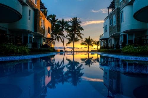 A serene tropical resort pool at sunset reflects palm trees and modern villas, creating a tranquil atmosphere ideal for vacation relaxation.
