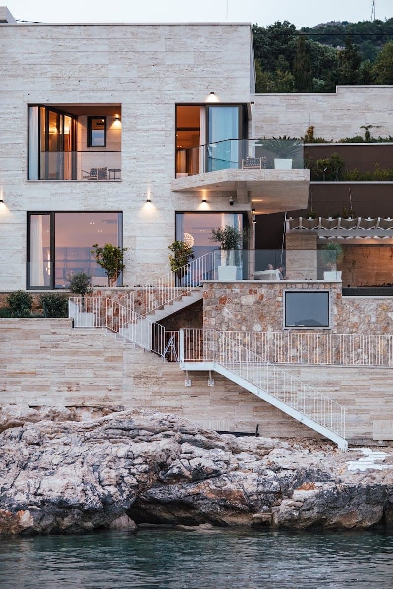 A luxurious modern villa perched on rocky cliffs overlooking the sea in Zaton, Croatia, exudes elegance with its stone facade, glass balconies, and outdoor seating areas, ideal for travelers seeking a serene coastal retreat.