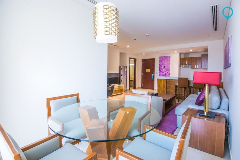 A cozy apartment interior features a modern dining area with a glass table, stylish chairs, and a warm pendant light, complemented by a vibrant living space with purple accents, ideal for travelers seeking comfortable accommodations.