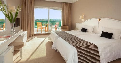 A cozy hotel room with a neatly made bed, balcony overlooking greenery, and natural light streaming in, ideal for travelers seeking relaxation and comfort.