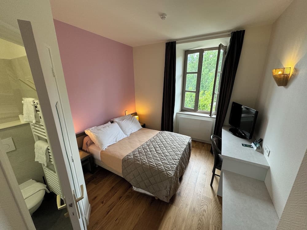 A cozy hotel room with a neatly made bed, wooden floors, a desk with a TV, and a view of greenery through an open window, ideal for travelers seeking comfort and tranquility.