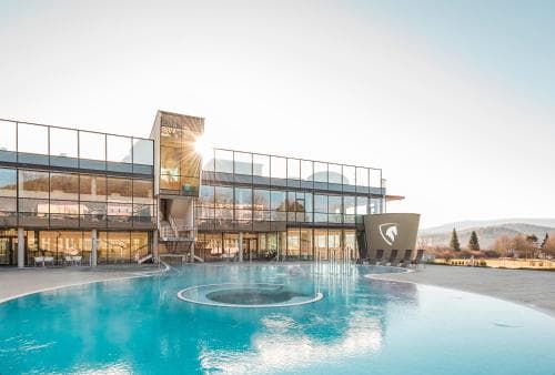 A serene outdoor thermal spa features a modern glass building with a sunlit pool, ideal for relaxation amidst nature, perfect for wellness retreats.