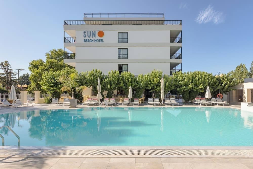 A serene beach hotel scene features a sparkling pool surrounded by lounge chairs and umbrellas, set against lush greenery and a modern white building under a clear blue sky, ideal for travelers seeking relaxation and leisure.