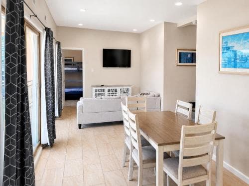 A cozy, well-lit vacation rental living room features a dining table, comfortable seating, a mounted TV, and bunk beds in the background, ideal for family getaways seeking comfort and convenience.