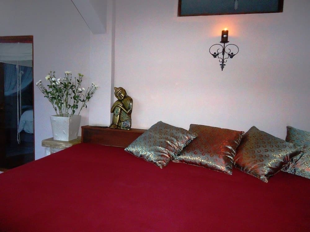 A serene bedroom features a cozy bed adorned with red linens and patterned pillows, complemented by a Buddha statue, fresh flowers, and a softly glowing wall sconce, ideal for travelers seeking tranquility and cultural ambiance.