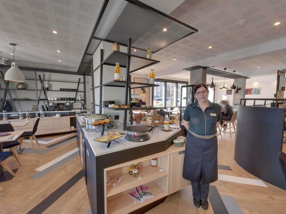 A welcoming café scene features a staff member standing by a well-stocked buffet counter with pastries and beverages, set in a modern, cozy interior with wooden floors and stylish shelving, ideal for travelers seeking a comfortable dining spot.
