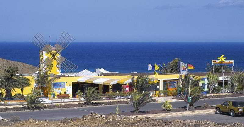 A vibrant coastal restaurant with a striking yellow windmill facade overlooks the serene ocean, surrounded by palm trees and flags, ideal for travelers seeking a picturesque dining spot near the sea.