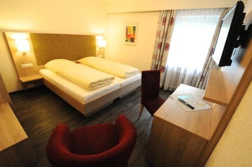 A cozy hotel room features a neatly made bed, warm lighting, a red armchair, and a desk by the window, creating an inviting atmosphere ideal for travelers seeking comfort and relaxation.