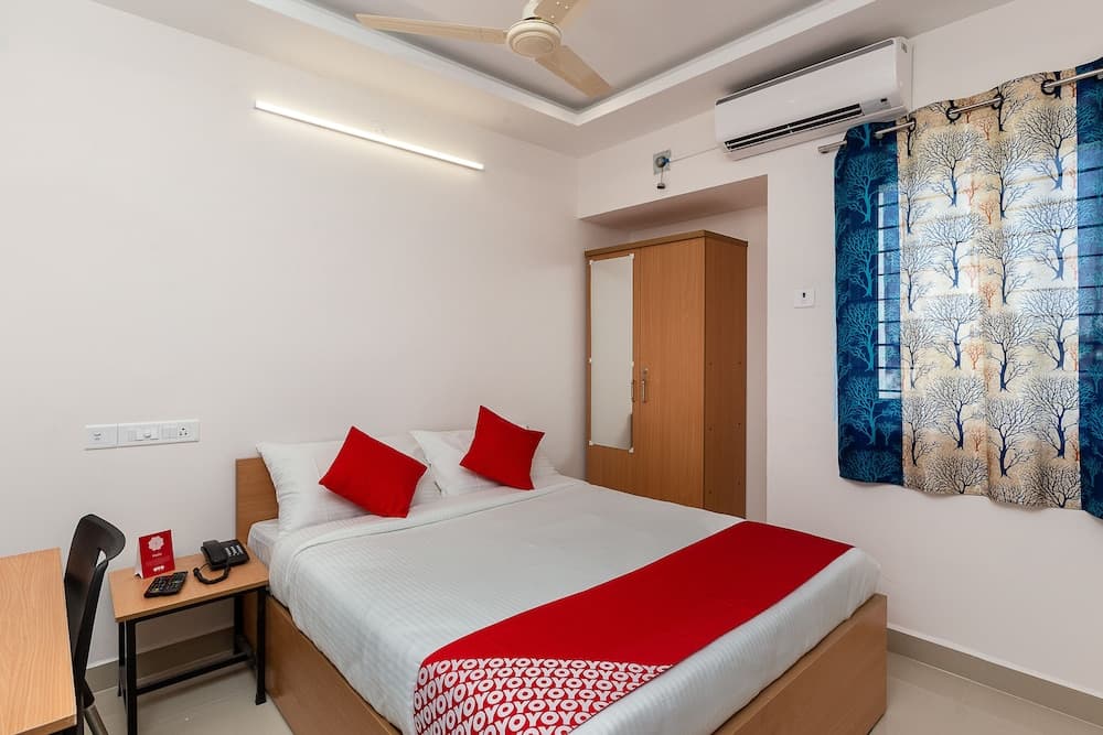 A cozy hotel room features a neatly made bed with red accents, wooden furniture, air conditioning, and tree-patterned curtains, ideal for travelers seeking comfort and convenience.