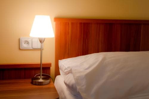 A cozy hotel room corner features a warmly lit bedside lamp on a wooden nightstand beside a neatly made bed, ideal for travelers seeking comfort and relaxation.