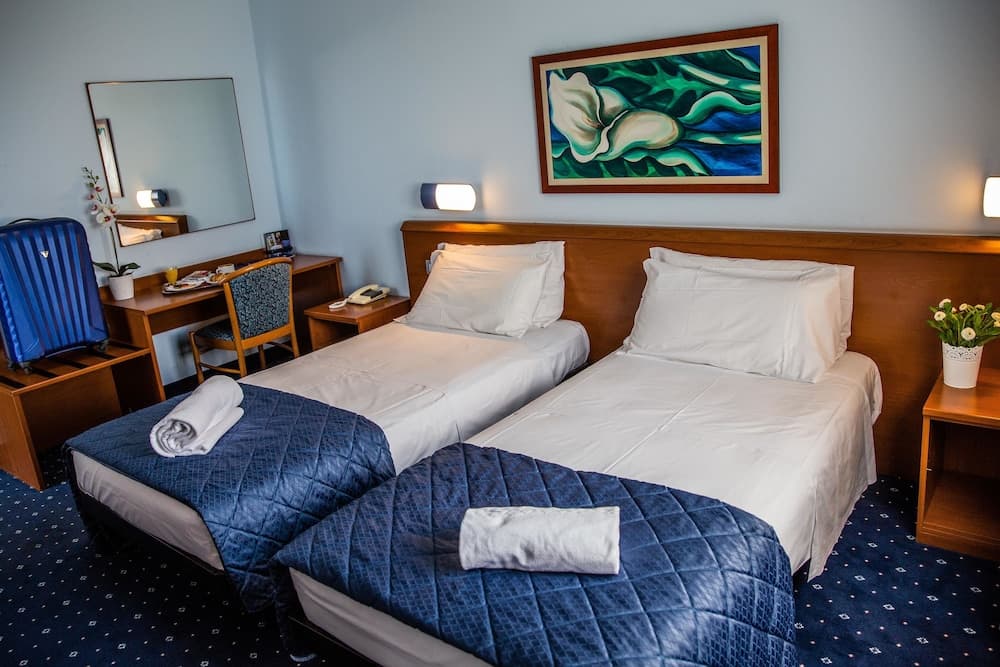 A cozy hotel room features two neatly made beds with blue quilts, a vibrant floral painting above the headboard, and a desk area with a mirror, ideal for travelers seeking comfort and convenience.