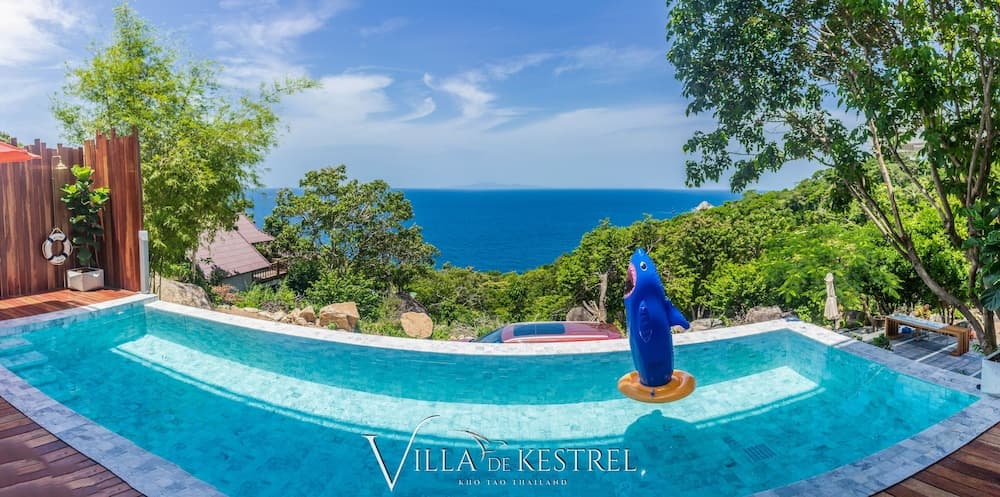 A serene tropical villa features a vibrant pool with a playful shark float, surrounded by lush greenery and overlooking the vast blue ocean, ideal for a luxurious beachside getaway.