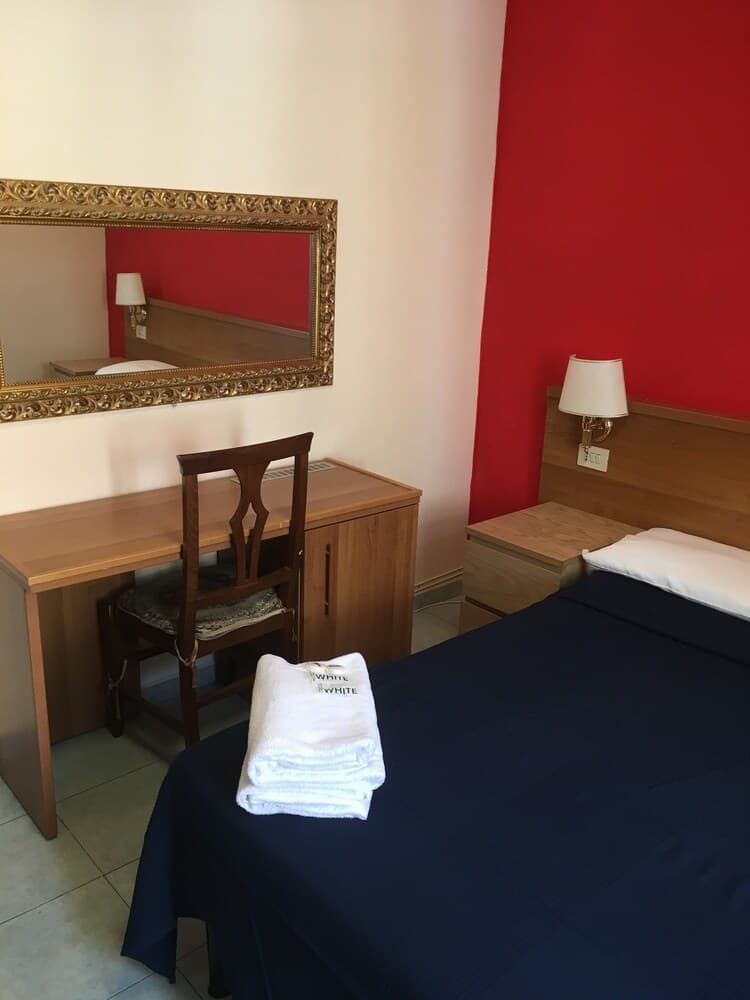 A cozy hotel room features a neatly made bed with navy blue linens, a wooden desk with a chair under an ornate mirror, and a vibrant red accent wall, ideal for travelers seeking comfort and simplicity.