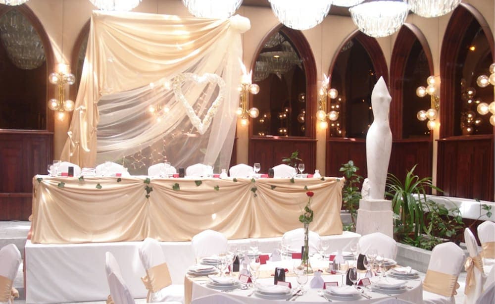 A beautifully decorated banquet hall with arched windows, elegant tables draped in gold, twinkling lights, and a heart-shaped backdrop creates a romantic atmosphere ideal for wedding receptions or special events.