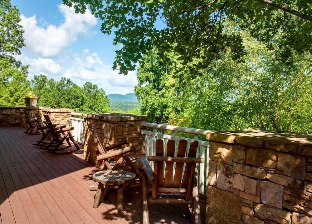 A serene wooden deck with rustic chairs and stone railings offers a tranquil retreat amidst lush greenery and distant mountain views, perfect for a relaxing getaway.