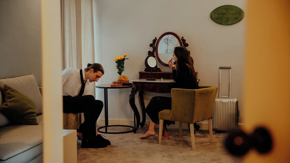A cozy hotel room scene captures a couple preparing for an outing, with a man tying his shoes by a sofa and a woman applying makeup at a vintage vanity adorned with flowers, evoking a warm and intimate travel moment.