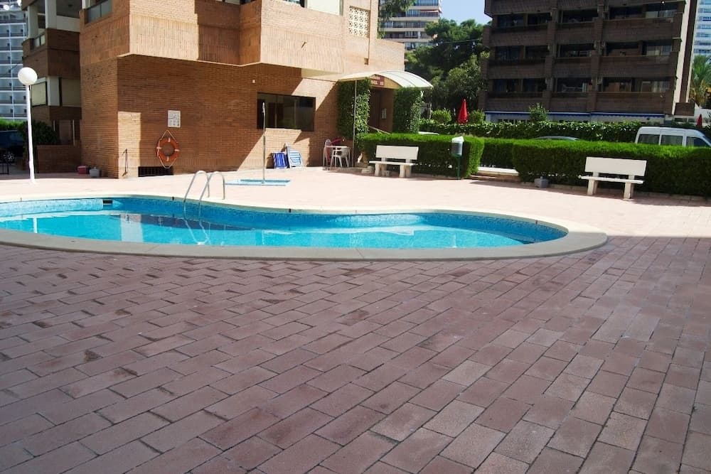 A serene urban resort pool area features a clear blue pool surrounded by brick pavement, benches, and lush greenery, ideal for travelers seeking relaxation in a cityscape setting.