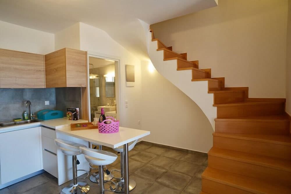 A cozy apartment interior features a modern kitchen with wooden cabinets, a small dining area with white stools, and a curved wooden staircase leading upstairs, ideal for travelers seeking compact yet stylish accommodations.