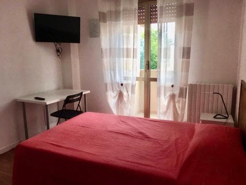 A cozy bedroom with a red bedspread, a desk by the window with sheer curtains, and a mounted TV, ideal for travelers seeking a comfortable and functional stay in a quiet setting.