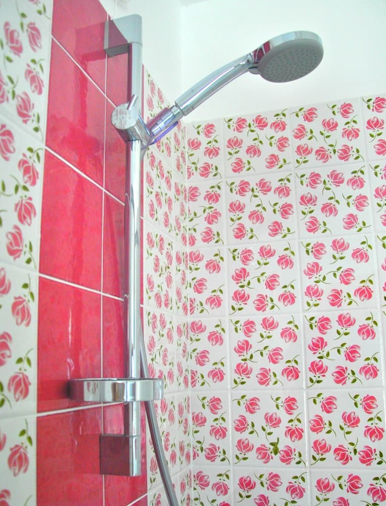 A vibrant bathroom corner features pink rose-patterned tiles and a sleek chrome showerhead, creating a cheerful and inviting atmosphere ideal for travelers seeking a cozy accommodation experience.