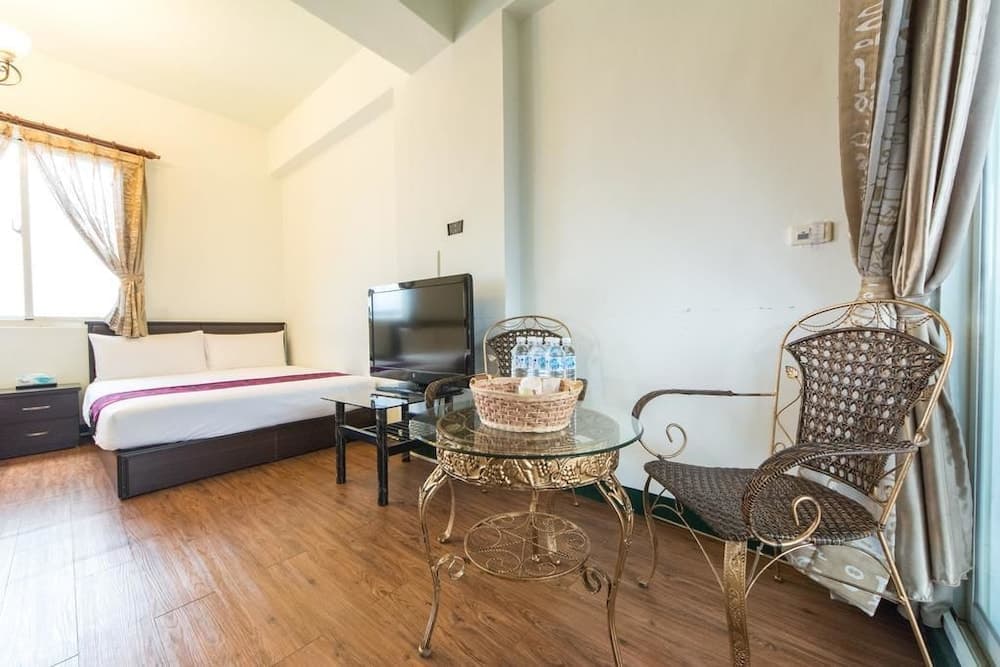 A cozy hotel room with wooden flooring, a neatly made bed, a glass-top table with wicker chairs, and natural light streaming through sheer curtains, ideal for travelers seeking comfort and relaxation.