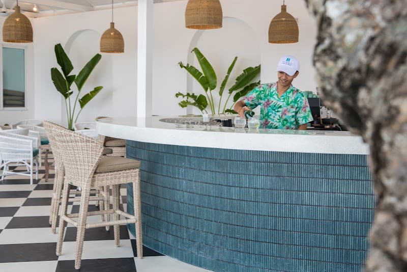 A bartender serves drinks at the stylish bar of Tembo Beach Club & Resort in Bangrak Beach, Thailand, featuring a vibrant tropical vibe with wicker chairs, lush plants, and woven pendant lights, perfect for travelers seeking a relaxing beachside retreat.