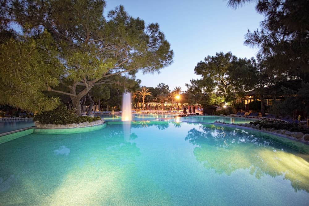 A serene evening at a luxurious resort features a glowing pool illuminated by a central fountain, surrounded by lush trees and ambient lighting, ideal for travelers seeking relaxation and tranquility.