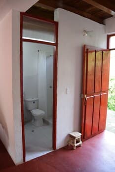 A cozy indoor space with a red-tiled floor, white walls, a wooden door leading outside, and an open doorway to a bathroom, evokes a rustic charm ideal for travelers seeking a quaint and serene accommodation experience.