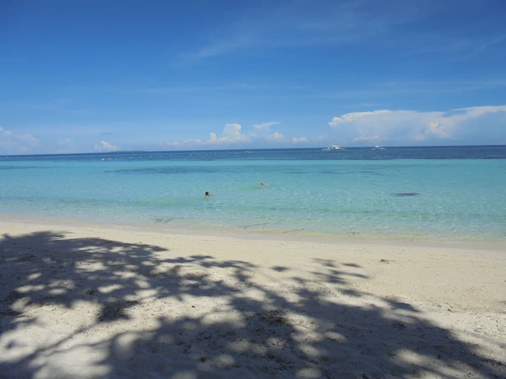 A serene tropical beach scene features clear turquoise waters where swimmers enjoy the calm sea under a bright blue sky, with tree shadows on the sandy shore, ideal for relaxation and vacation photography.