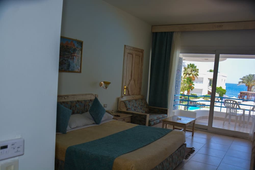 A cozy hotel room with a neatly made bed, wooden furnishings, and a sliding glass door leading to a balcony overlooking a serene beachfront, perfect for a relaxing seaside getaway.