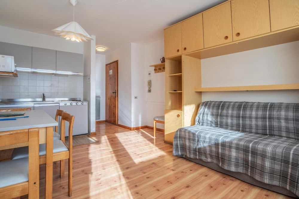 A cozy studio apartment with wooden flooring, a plaid-covered bed, and a compact kitchen area bathed in natural light, ideal for travelers seeking a comfortable and functional short-term stay.