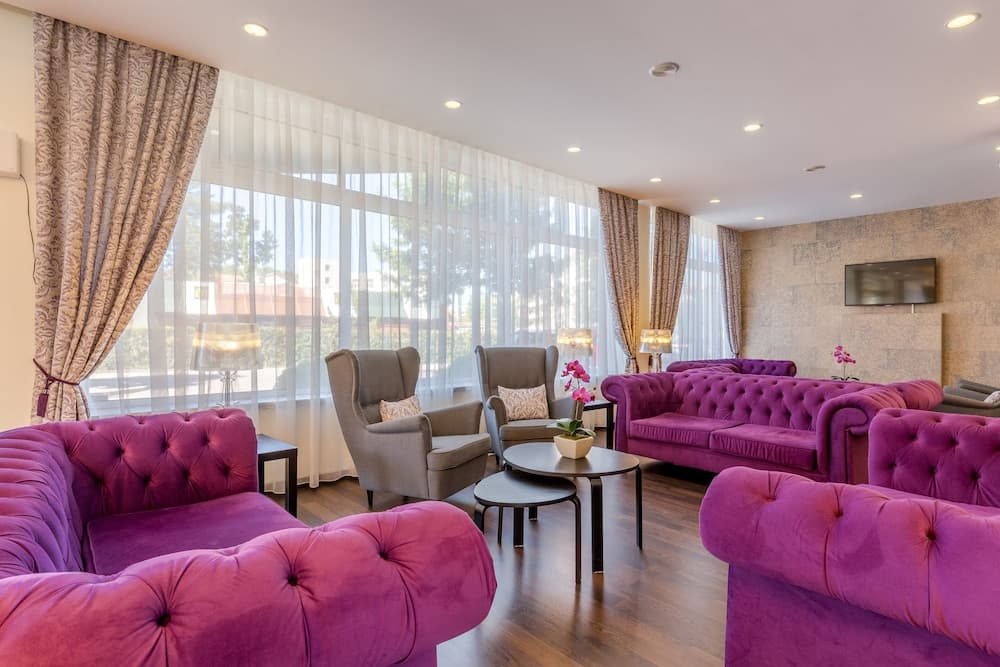 A cozy hotel lounge features plush purple sofas, elegant gray armchairs, and large windows with sheer curtains, creating a welcoming atmosphere ideal for relaxation or business meetings.
