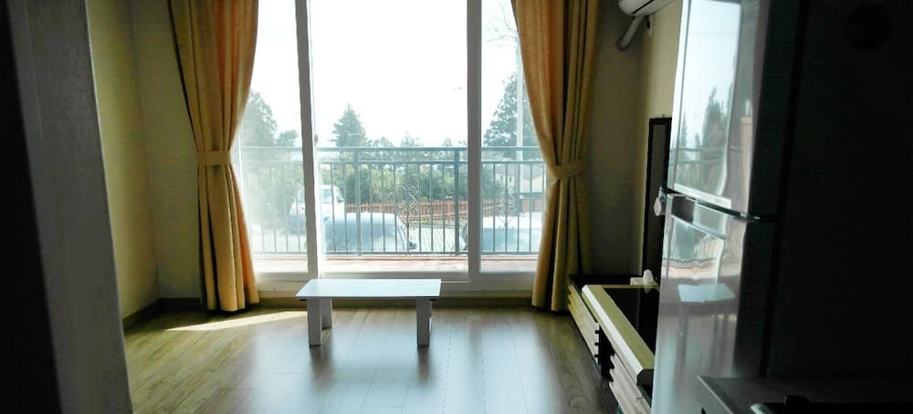 A cozy apartment interior features a minimalist setup with a small table, wooden flooring, and large windows draped with golden curtains, offering a serene view of greenery outside, ideal for travelers seeking a peaceful urban retreat.