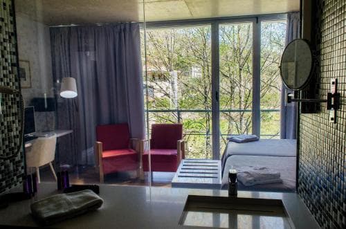 A cozy hotel room with vibrant red chairs, a glass door leading to a tree-lined balcony, and tiled walls creates a serene and inviting atmosphere, perfect for travelers seeking relaxation and comfort.