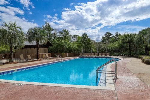 A serene outdoor pool surrounded by lush greenery under a partly cloudy sky offers a tranquil retreat, ideal for vacation relaxation with lounge chairs and a wooden cabana nearby.