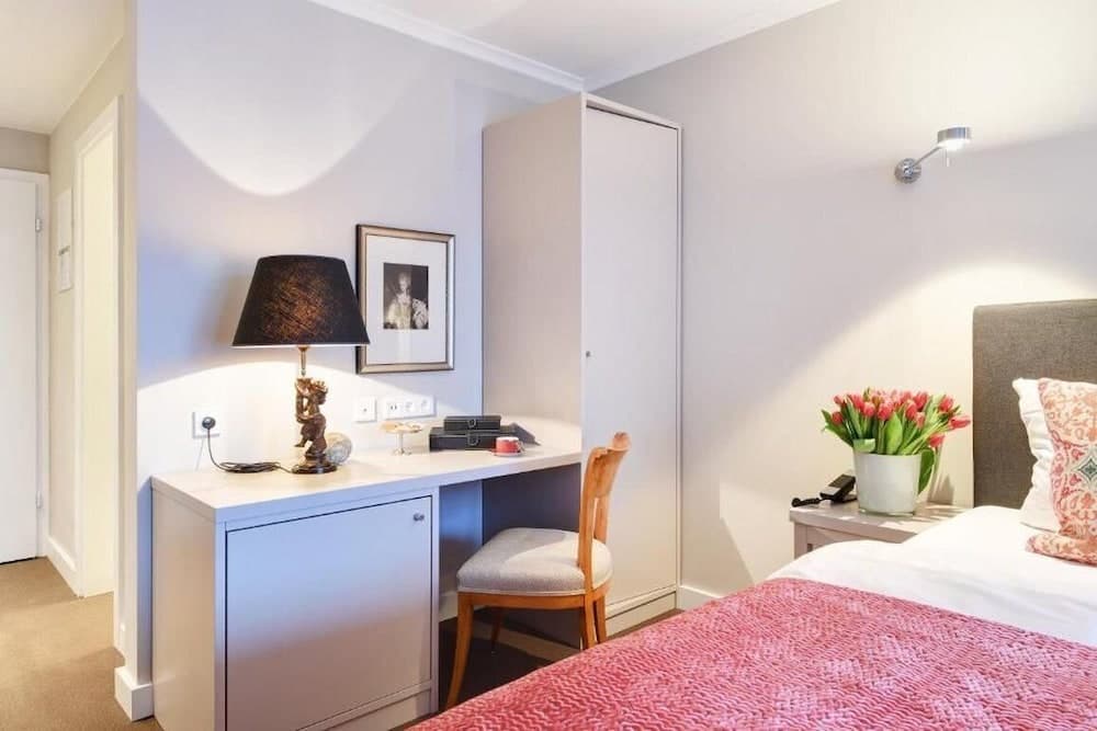 A cozy hotel room features a neatly made bed with a pink quilt, a desk adorned with a lamp and framed portrait, vibrant tulips on the bedside table, and a serene ambiance ideal for travelers seeking comfort and relaxation.