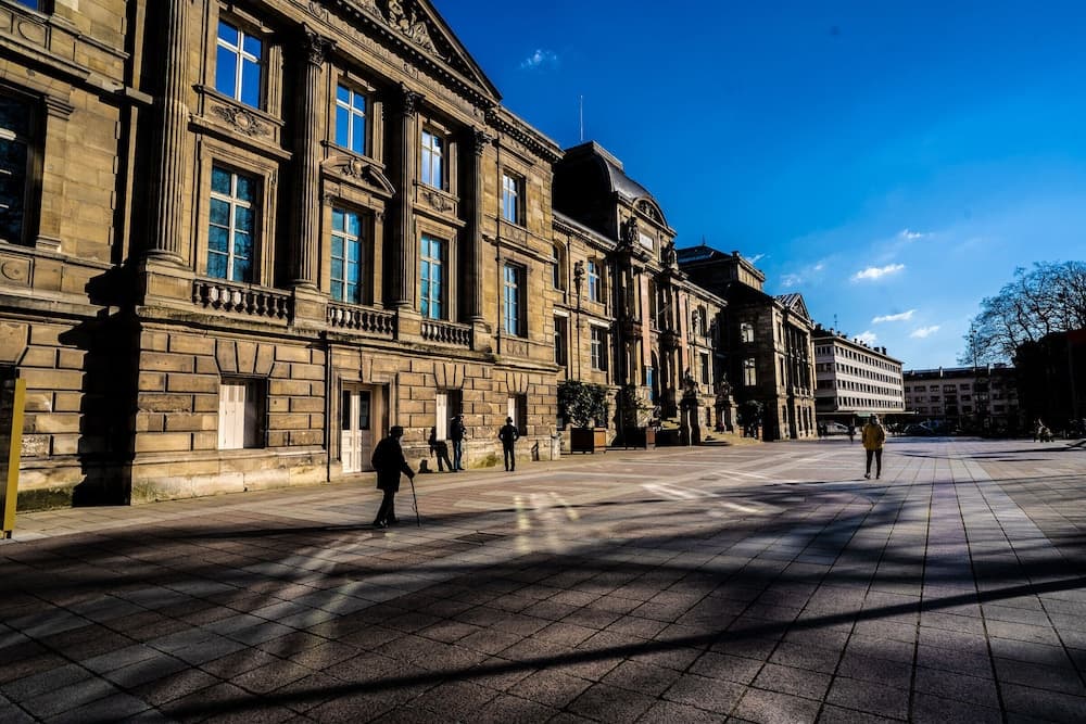 A serene European square bathed in sunlight features grand historic architecture with ornate facades, casting long shadows on the cobblestone ground, ideal for cultural exploration and photography.