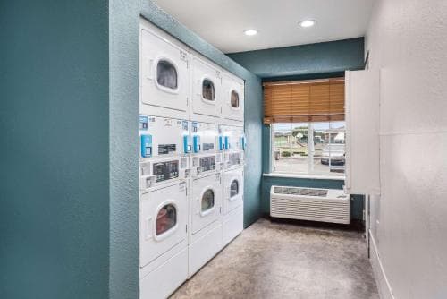 A clean, well-lit laundry room with multiple washing machines and dryers, featuring teal walls, a window with blinds, and an air conditioning unit, offers a convenient facility for travelers needing to wash clothes during their stay.