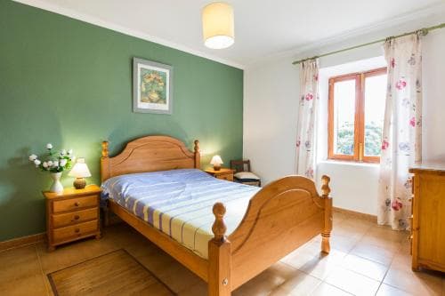 A cozy bedroom with a wooden bed, green walls, floral curtains, and natural light streaming through the window, ideal for travelers seeking a serene and comfortable accommodation.