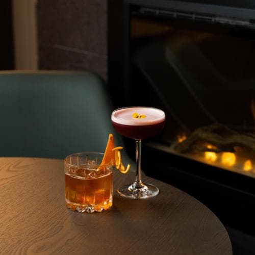 A cozy lounge setting features two elegantly garnished cocktails on a wooden table beside a glowing fireplace, evoking warmth and sophistication ideal for a romantic evening out.