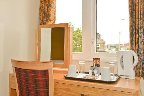 A cozy hotel room features a wooden vanity with a mirror, a tray of tea-making essentials, and a view through patterned curtains to a quiet street, ideal for travelers seeking comfort and convenience.