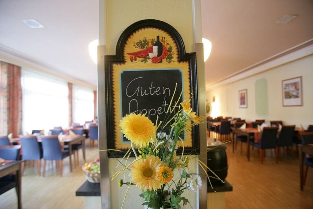 A cozy restaurant interior features a welcoming "Guten Appetit" sign adorned with vibrant yellow flowers, set against a backdrop of neatly arranged tables and chairs, ideal for travelers seeking a warm dining experience.